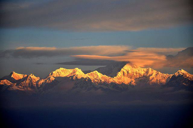 Snow-capped mountain range glowing orange and gold at sunset under a partly cloudy blue sky
